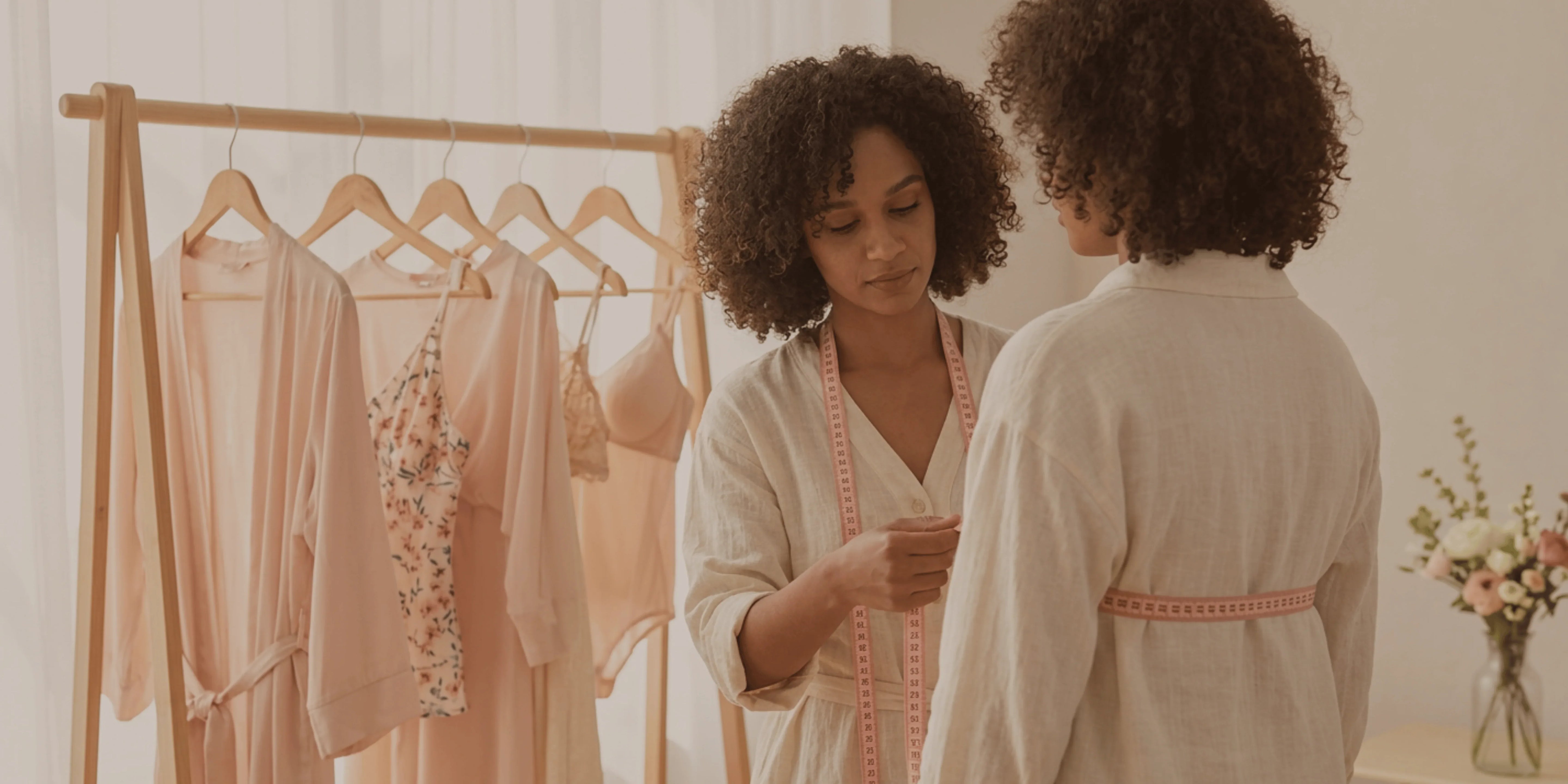 Two women in a room with a rack of pink dresses and a vase of flowers.